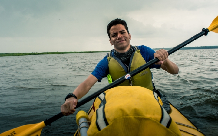 A young person smiles at the camera while paddling on a large body of water. 